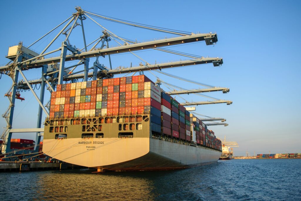 A large container ship at an industrial harbor with cranes under a clear blue sky.